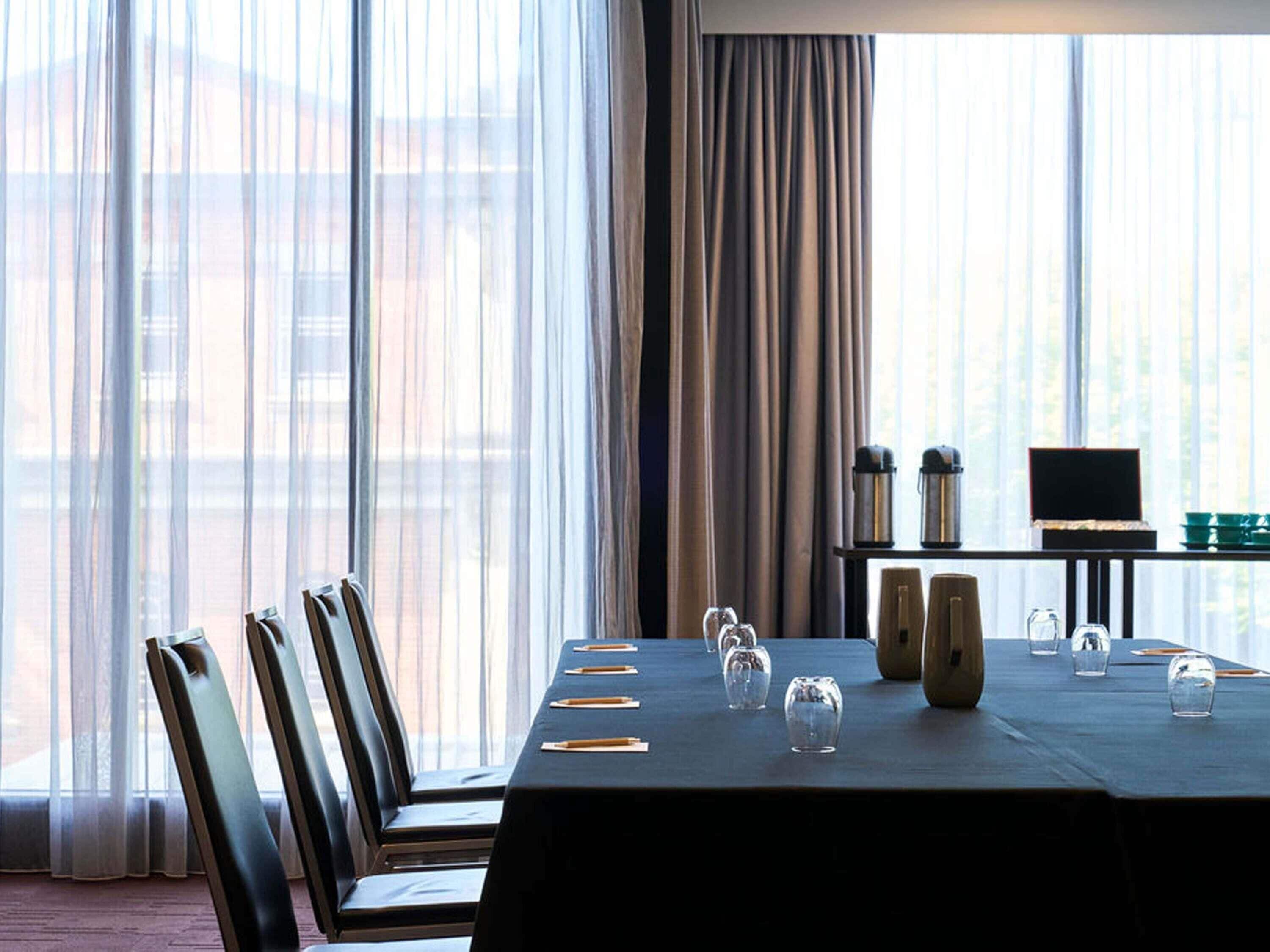 Habitación de hotel con una mesa de comedor preparada para cuatro personas, un televisor sobre un mueble bajo y vistas a través de amplias ventanas.