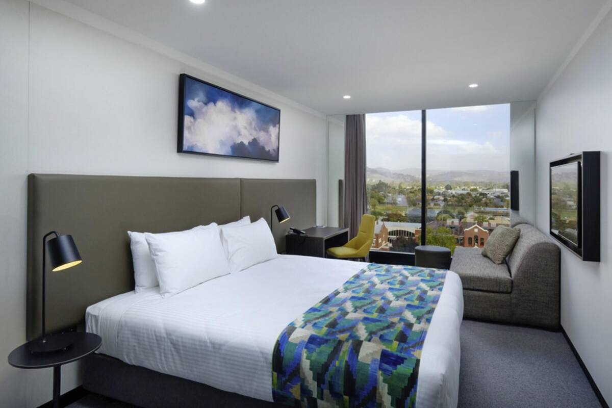 Une chambre d’hôtel moderne, dotée d’un grand lit, d’un bureau, d’une chaise et offrant une vue sur le paysage urbain.