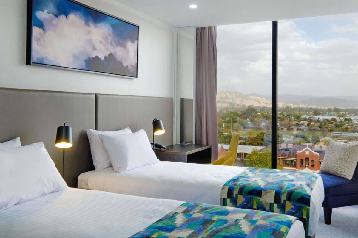 Une chambre d’hôtel avec deux lits, une grande fenêtre donnant sur la ville, un tableau au mur et une table de chevet avec une lampe.