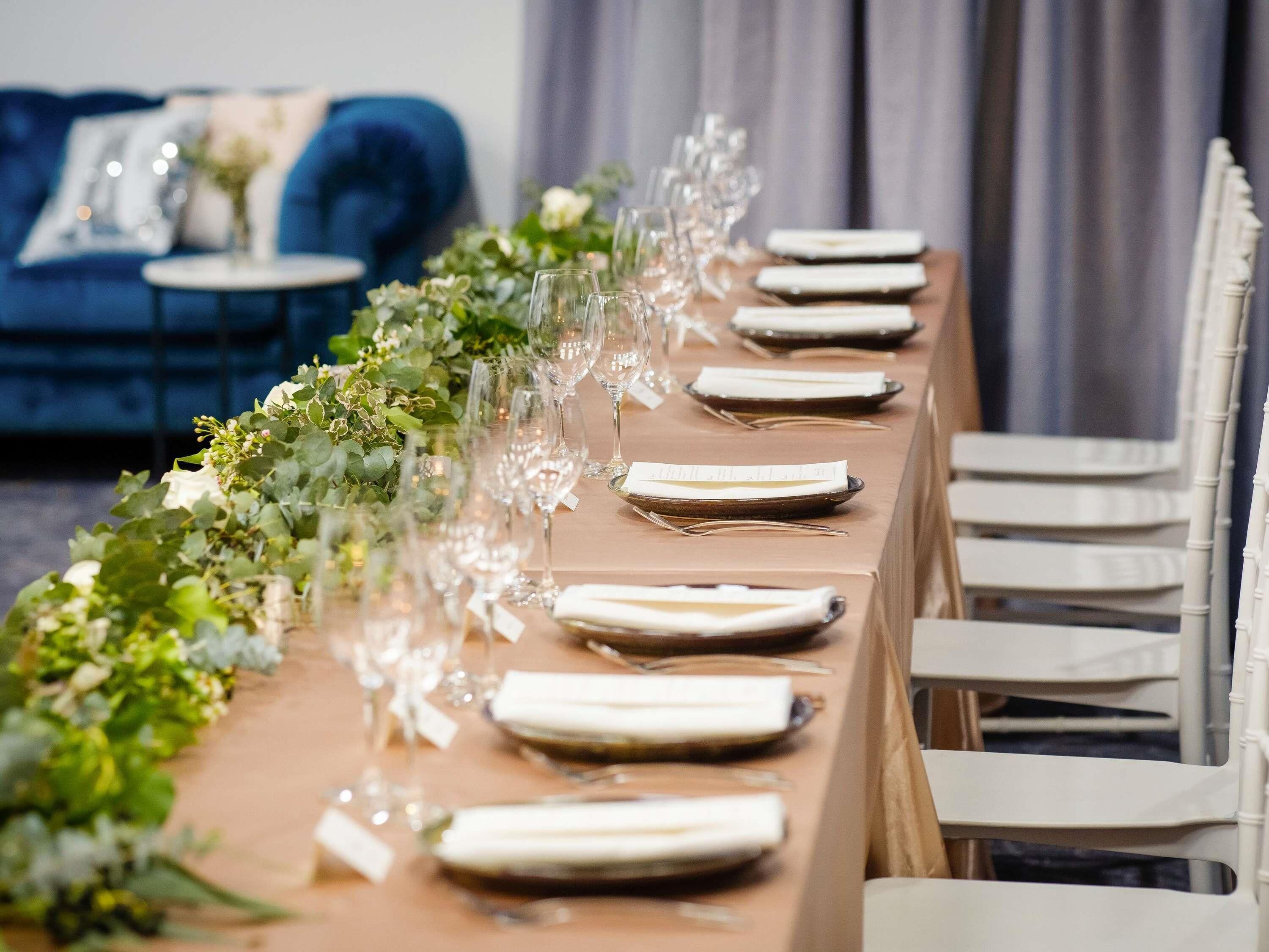 A long dining table with white chairs, set with plates, glasses, and a centerpiece of greenery.