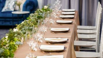 A long dining table with white chairs, set with plates, glasses, and a centerpiece of greenery.