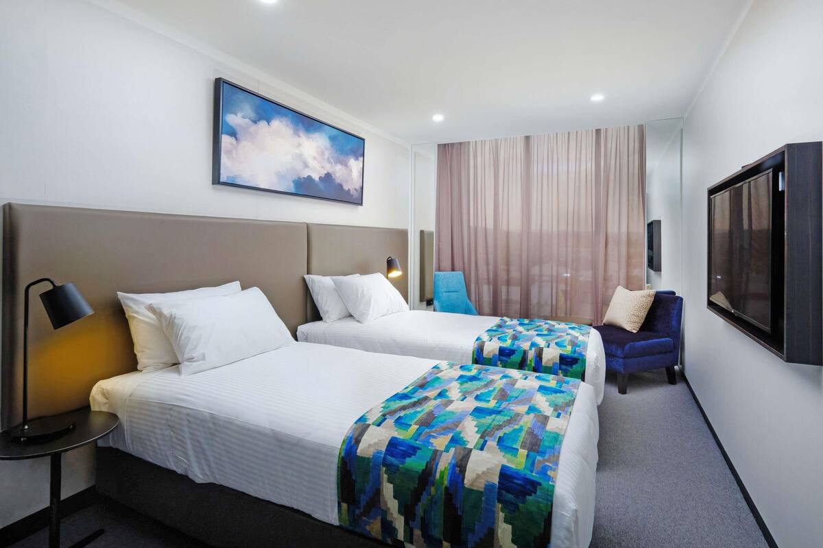 Une chambre d’hôtel avec deux lits, une télévision, un tableau et une fenêtre avec des rideaux.