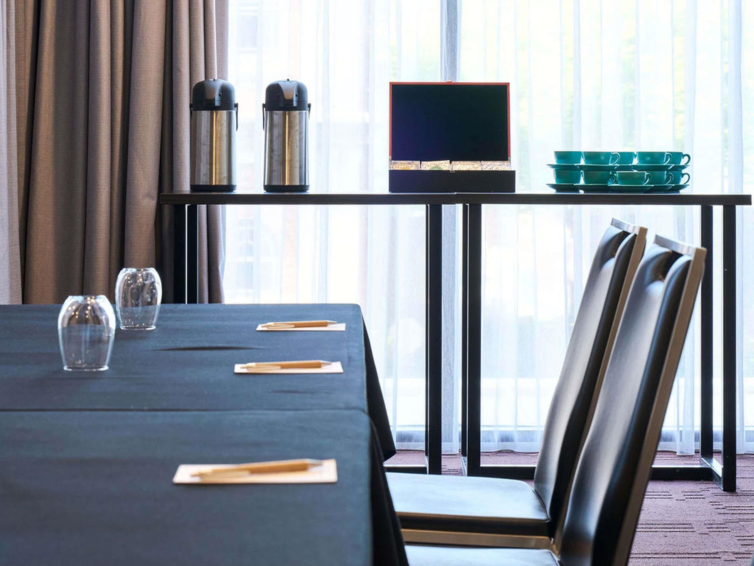 A hotel room with a dining table set for two, a laptop, two thermos flasks, a green tray, and a window with curtains.