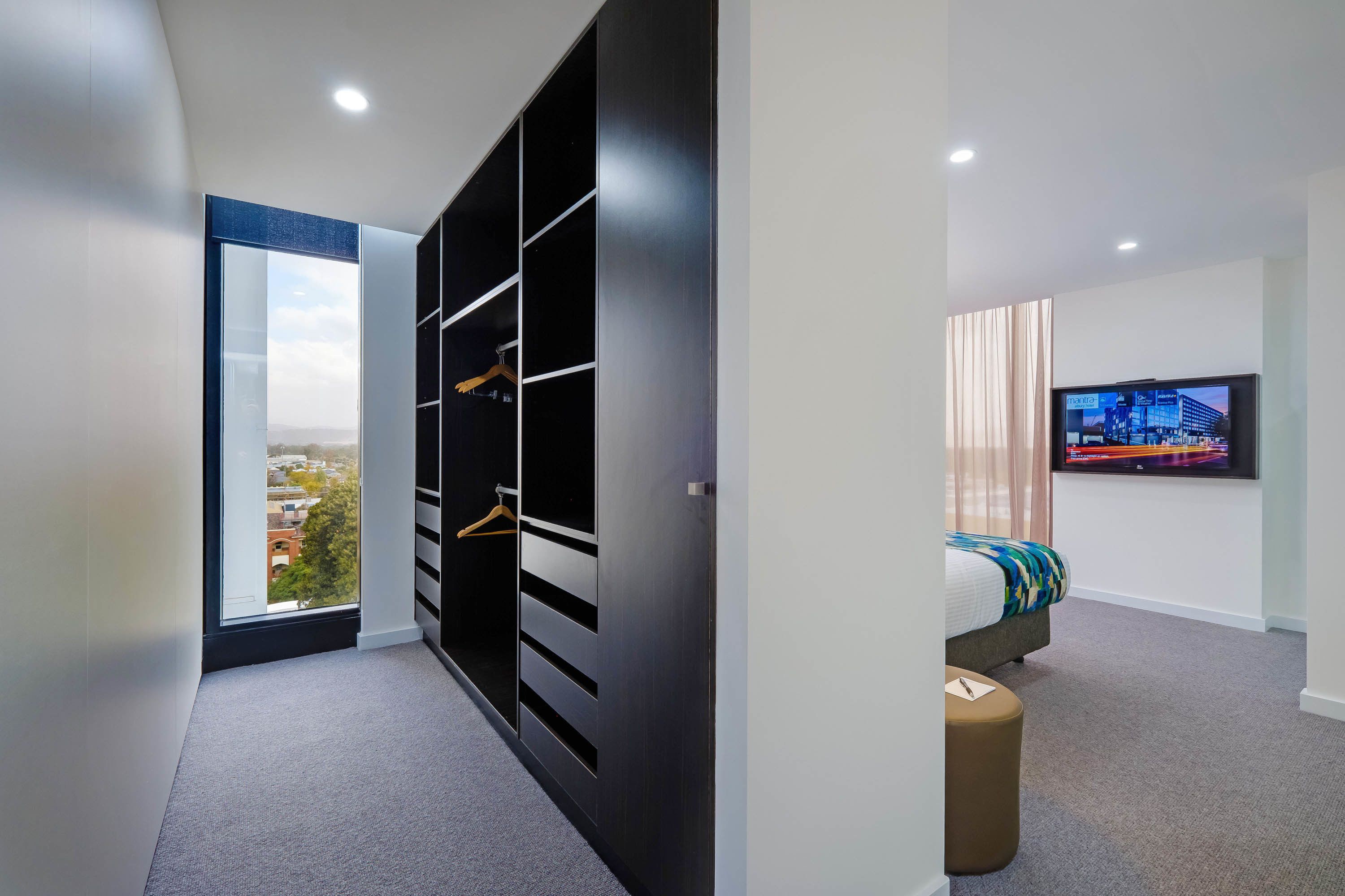 Una habitación de hotel moderna con un ventanal grande, un armario con varios compartimentos, una cama con una manta colorida y un televisor de pantalla plana montado en la pared.