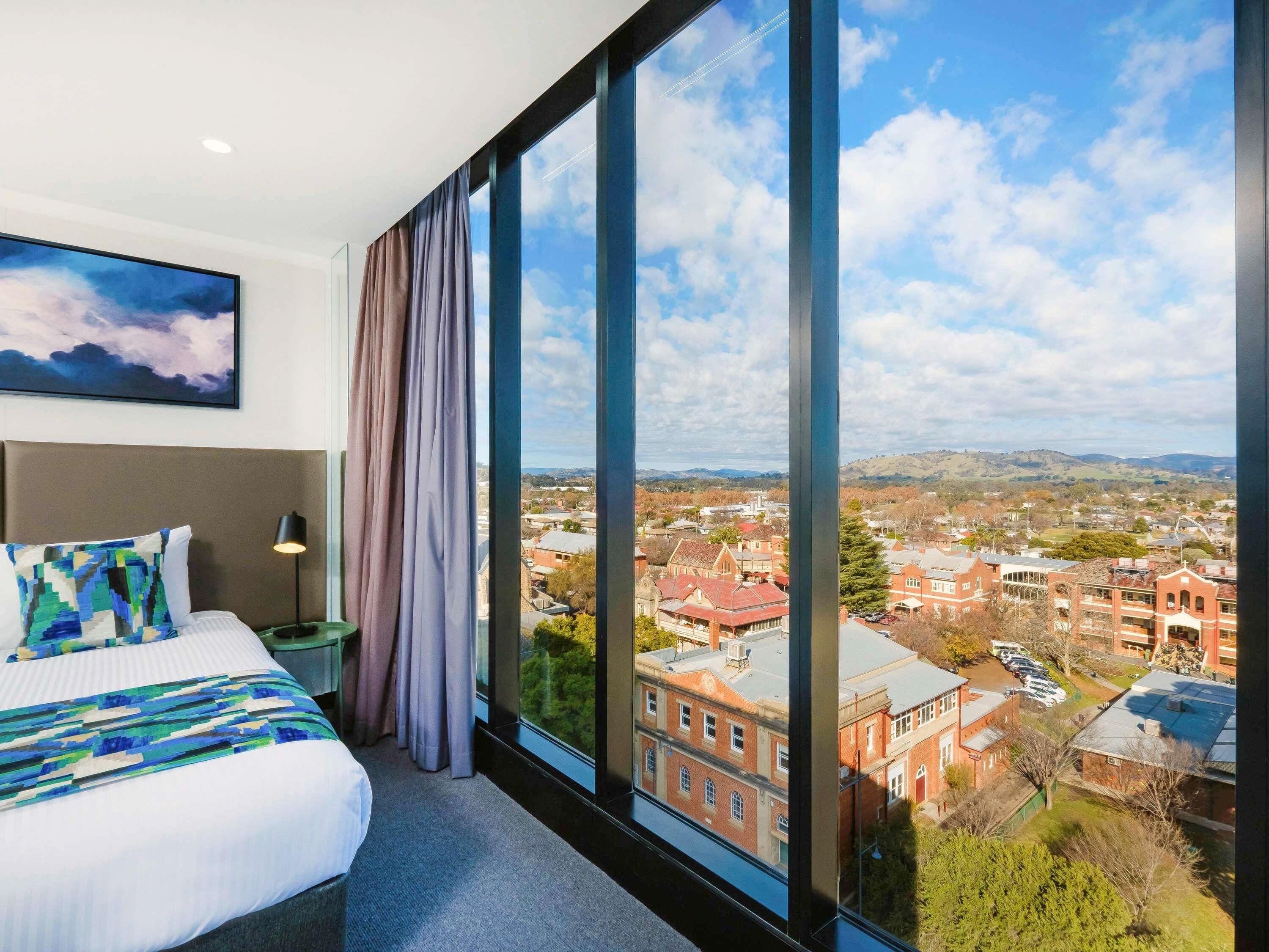 A hotel room with a large window, a bed, a nightstand, and a painting on the wall.