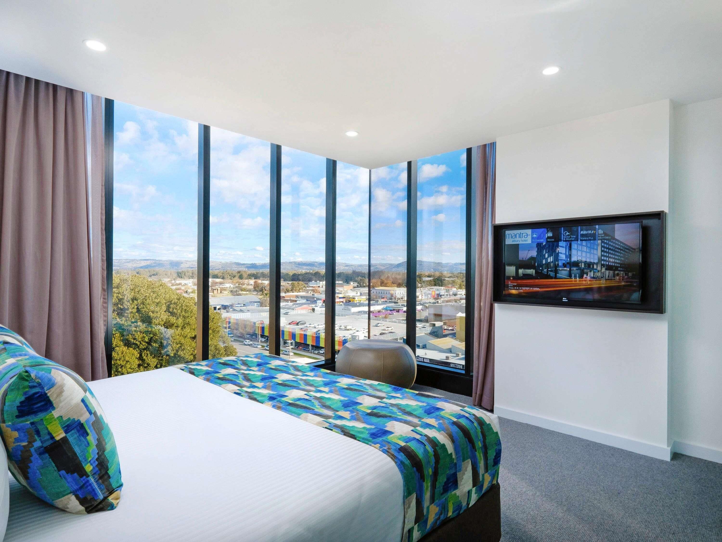 A hotel room with a large bed, a television, and a view of a cityscape through large windows.