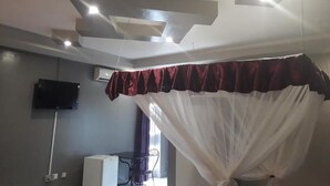 Une chambre à coucher avec un lit recouvert d’un fin baldaquin blanc, un téléviseur à écran plat fixé au mur et un appareil de climatisation blanc.