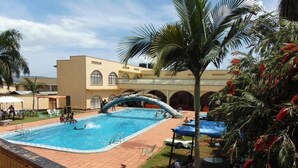 Une piscine avec un toboggan, entourée de palmiers et bordée d’un bâtiment aux fenêtres en arc de cercle.
