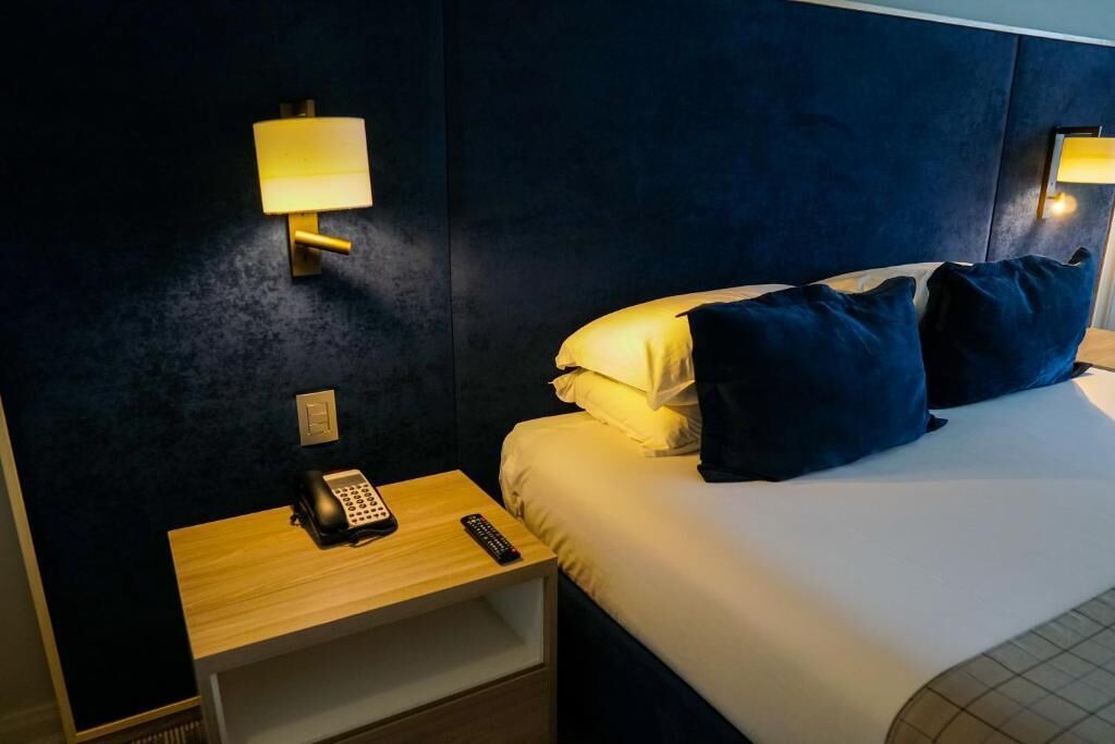 Une chambre d’hôtel avec un lit, deux lampes de chevet, une table de chevet avec un téléphone et une télécommande, et une tête de lit bleu foncé.