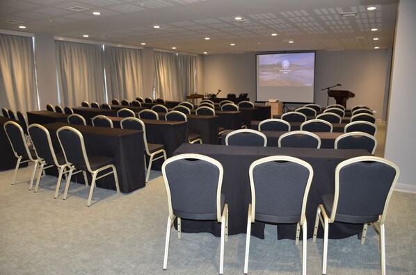 Un salón de conferencias con filas de sillas, una mesa grande, un atril y una pantalla para presentaciones.