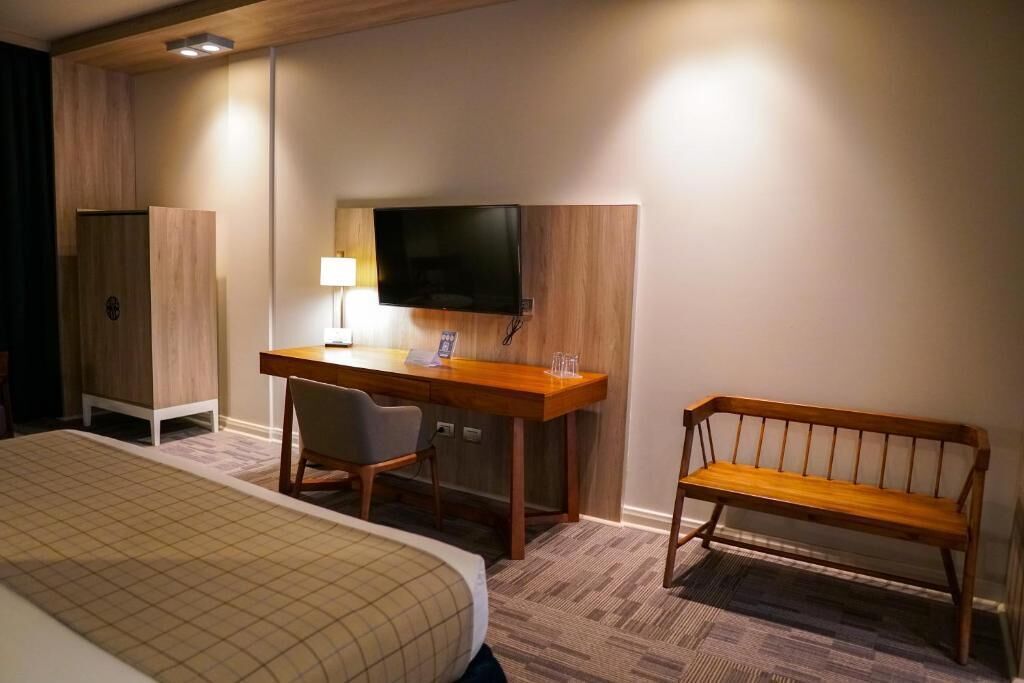 A hotel room with a bed, a desk with a chair, a television mounted on the wall, and a bench.