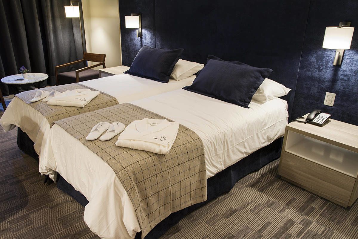 A hotel room with two beds, a bedside table with a lamp, a chair, and a small round table with a vase.