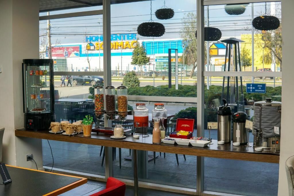 Una barra de petits dejUnis amb diferents aliments i condiments, una cafetera i una vista cap a una farmàcia i un aparcament a l'exterior.