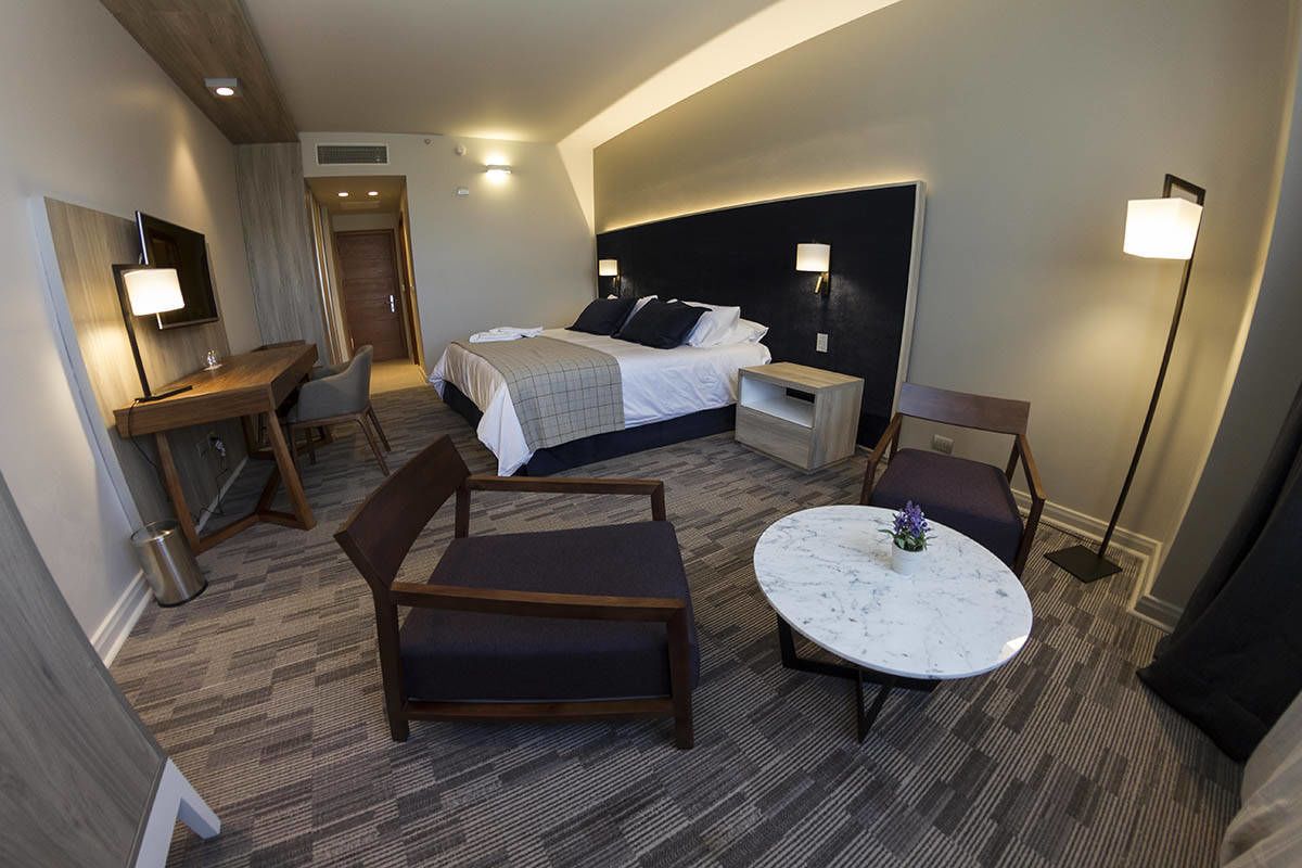 Habitación de hotel con una cama, dos mesitas de noche, un escritorio con una silla, una mesa de centro redonda y un televisor montado en la pared.