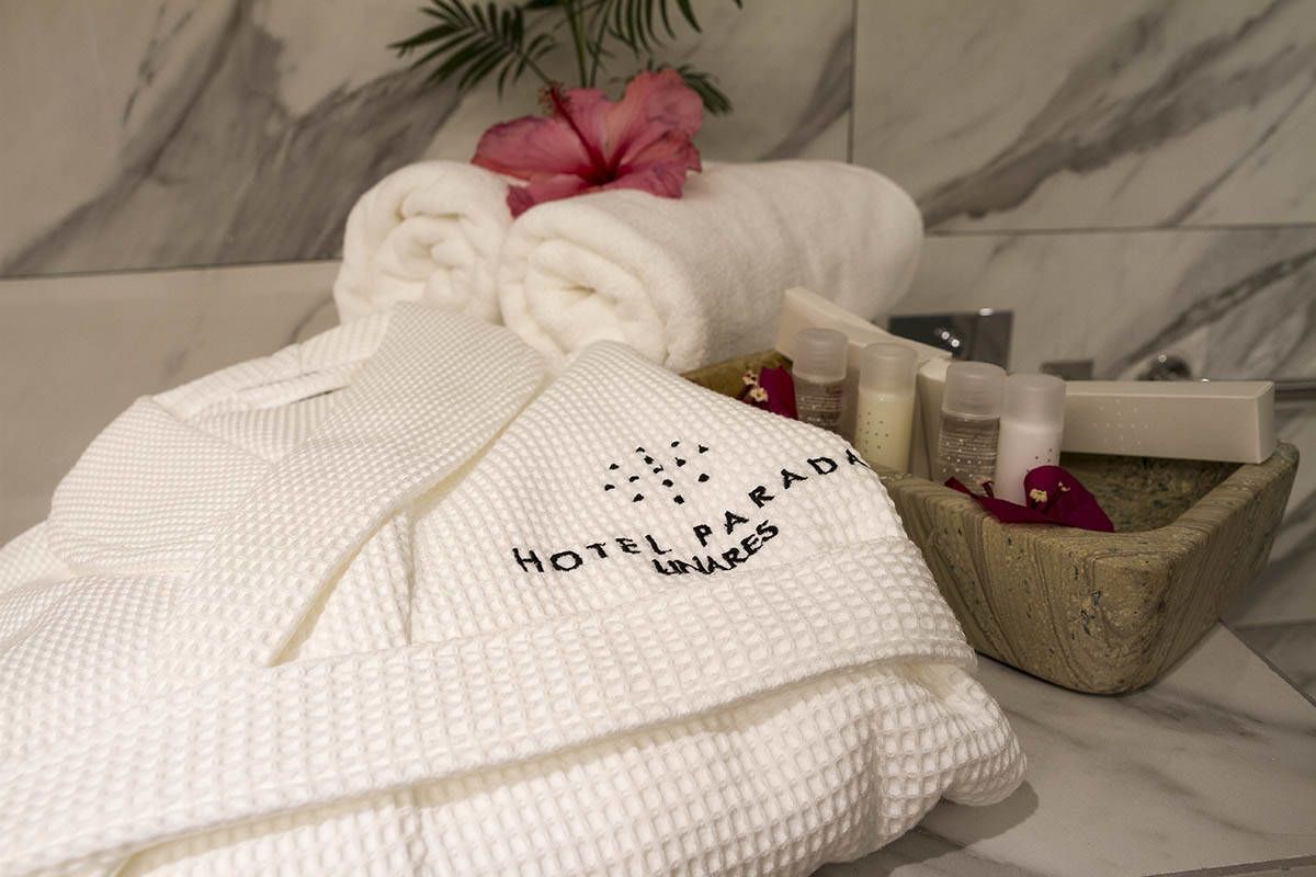 Une salle de bain bien aménagée, avec un mur en marbre, une serviette blanche portant l’inscription « HOTEL PARADA UNARES » et des articles de toilette rangés dans un récipient en bois.