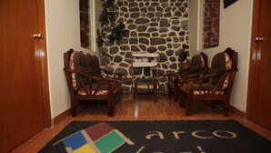 A lobby with two wooden chairs, a small table, a stone wall, and a doormat with a logo. - Marco Wasi - San Blas (Cusco)