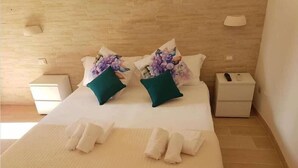 Un letto rifatto con cura, con cuscini a motivi floreali, una coperta bianca e asciugamani sul letto. Sono presenti due comodini con lampade e una piccola scatola su uno di essi.