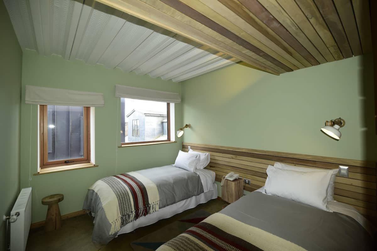 A bedroom with two beds, a wooden ceiling, and two windows with white blinds.