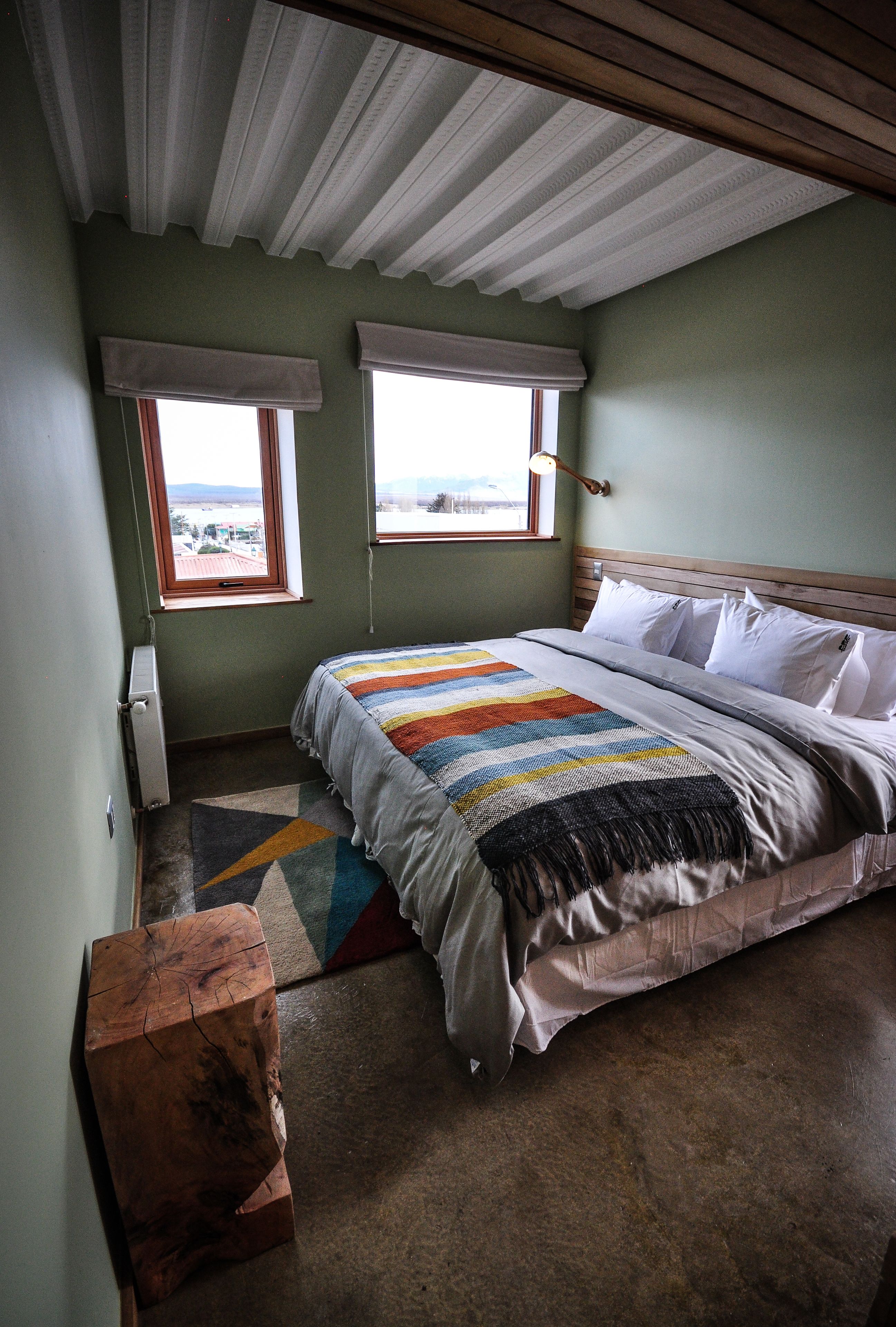 Un dormitorio con una cama grande, dos ventanas con persianas, un radiador y un banco de madera.