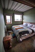Un dormitorio con una cama grande, dos ventanas con persianas, un radiador y un banco de madera.