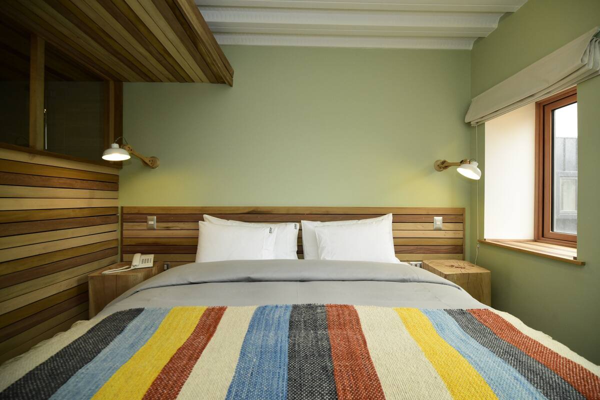 A bedroom with a wooden headboard, a striped bedspread, two bedside tables, and a window with a view.