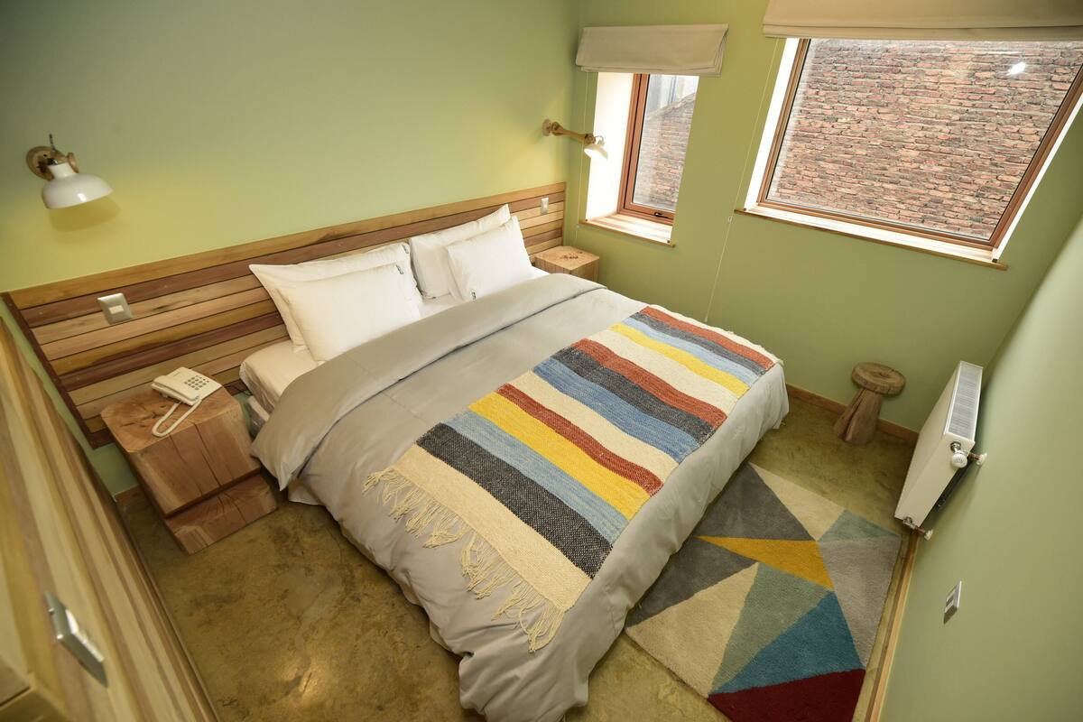 A bedroom with a wooden headboard, a striped bedspread, a bedside telephone, and a window with a view of a brick wall.