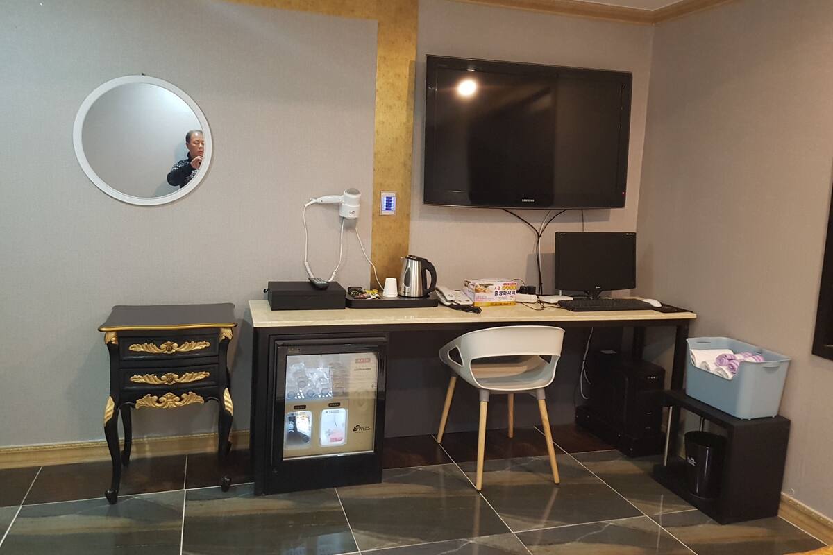 A compact hotel room with a desk, a chair, a TV, a small fridge, a mirror, and a trash bin.