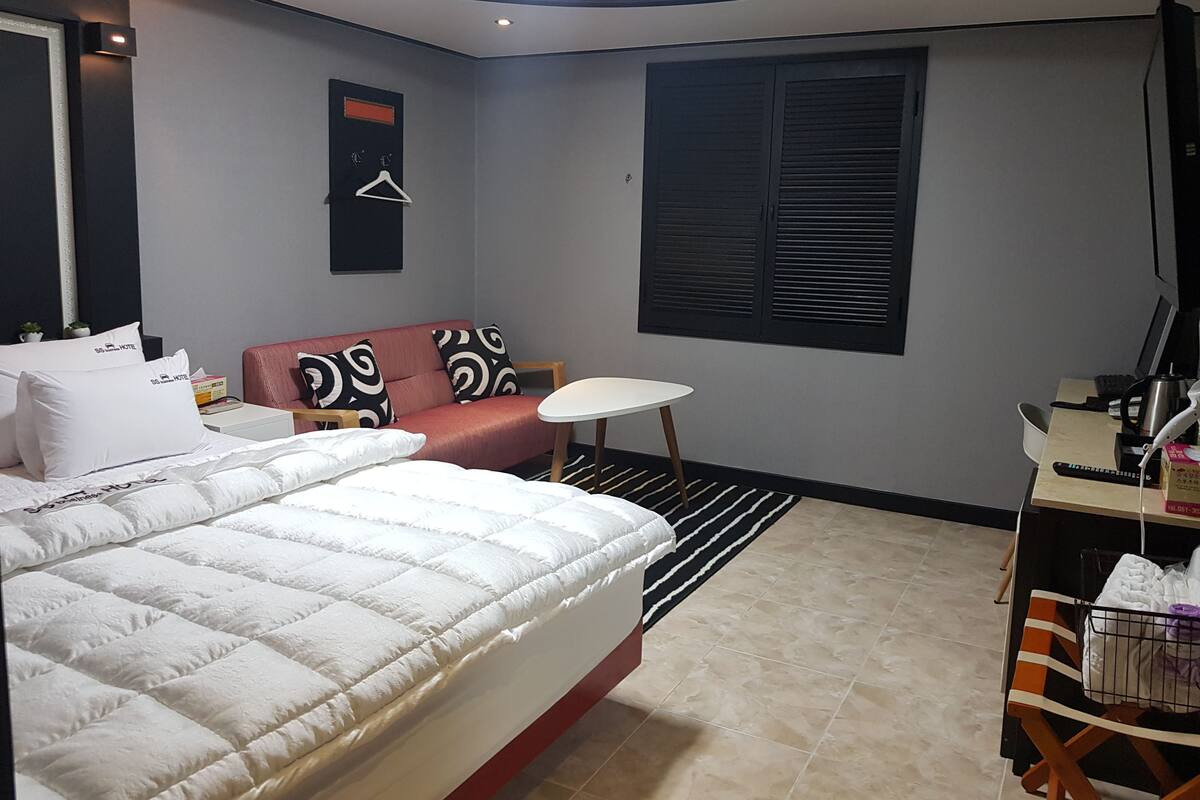 A hotel room with a bed, a sofa, a small table, a desk with a computer, and a window with shutters.