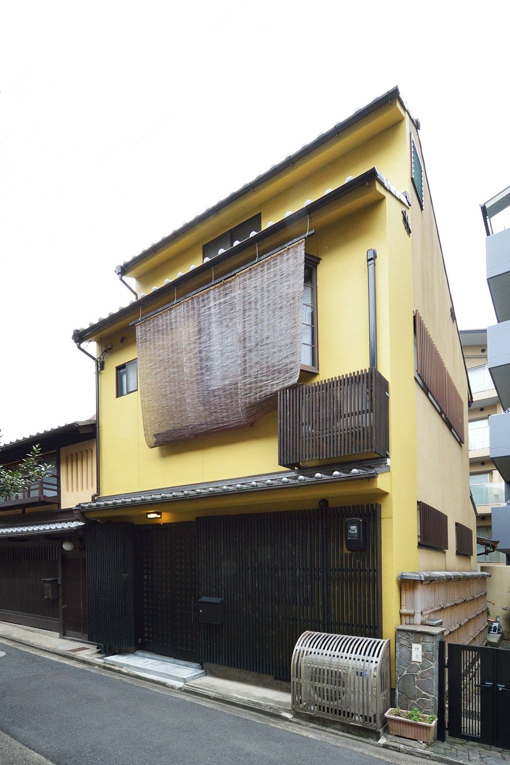 Um edifício de dois andares com fachada amarela, um portão preto grande e uma pequena construção de pedra na entrada.