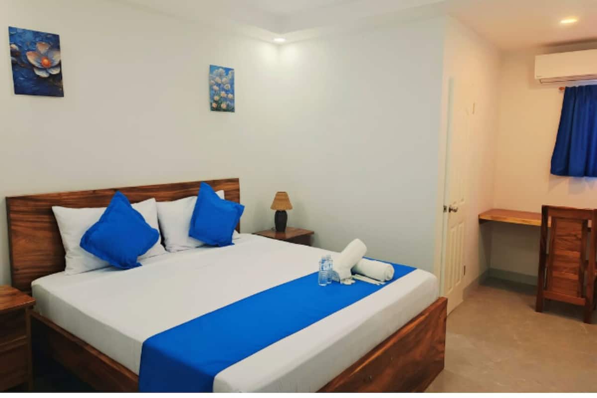 Una cama bien hecha con ropa de cama azul y blanca, cabecera de madera y corredor azul. Hay una mesita de noche con una lámpara y una pequeña mesa con una botella de agua y una toalla.