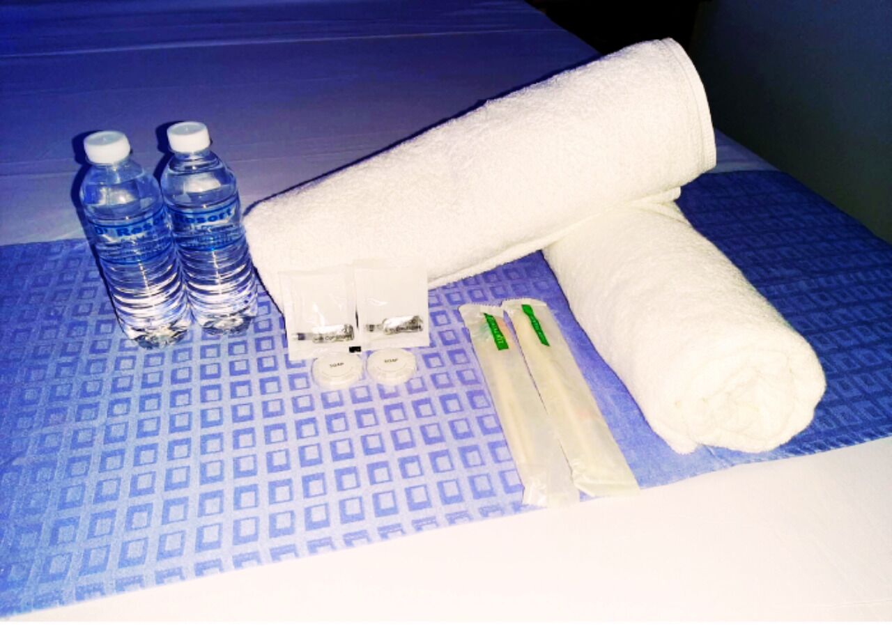 Una cama con una colcha azul estampada, dos botellas de agua, una toalla blanca y un par de cepillos de dientes verde y blanco.