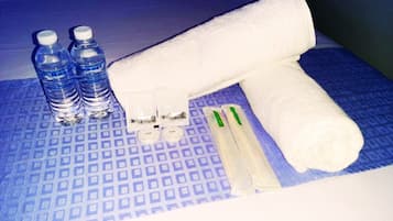 A bed with a blue patterned bedspread, two bottles of water, a white towel, and a pair of green and white toothbrushes.