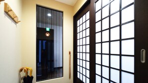 A hallway with a wooden door with a grid pattern, a window with vertical blinds, and a cane leaning against the wall. - Raku Inn (Kyoto)