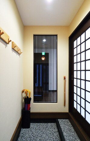 A hallway with a wooden floor, a black and white patterned mat, a black sliding door with a grid pattern, and a window with vertical blinds.