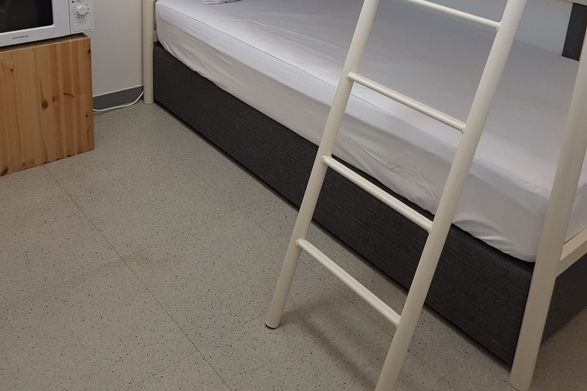 Una cama litera con escalera, un microondas y una tetera sobre un mueble de madera en una habitación con suelo de baldosas.