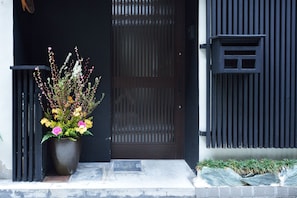 Una puerta negra con listones verticales, una planta en maceta con flores amarillas y rosadas, y una pequeña ventana a la derecha.