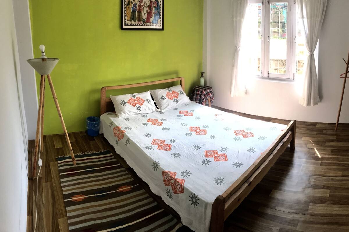 A bedroom with a bed, a standing lamp, a window with curtains, and a painting on the wall.