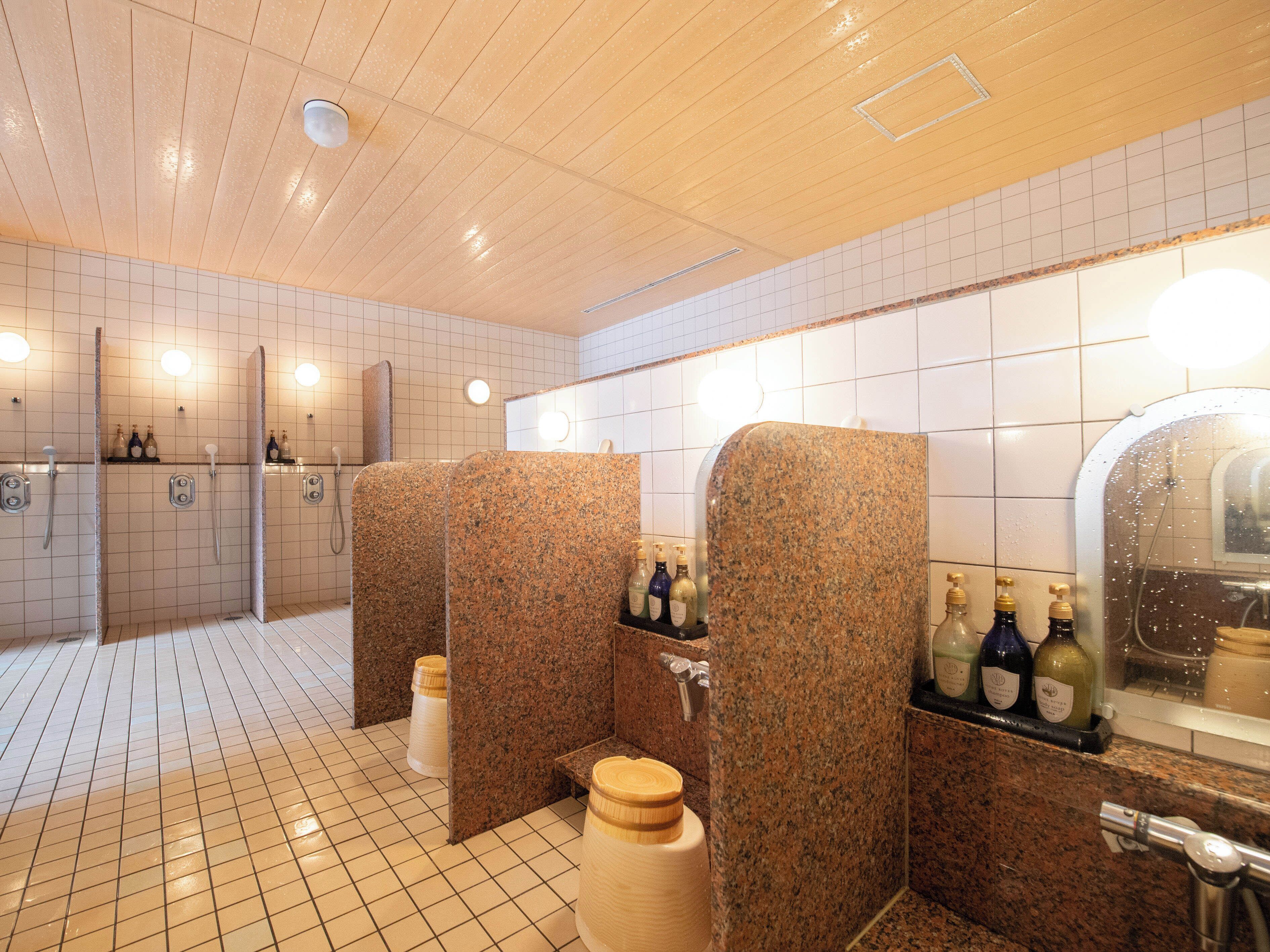 A public restroom with granite partitions, tiled floors, and a mirror with bottles of hand sanitizer.