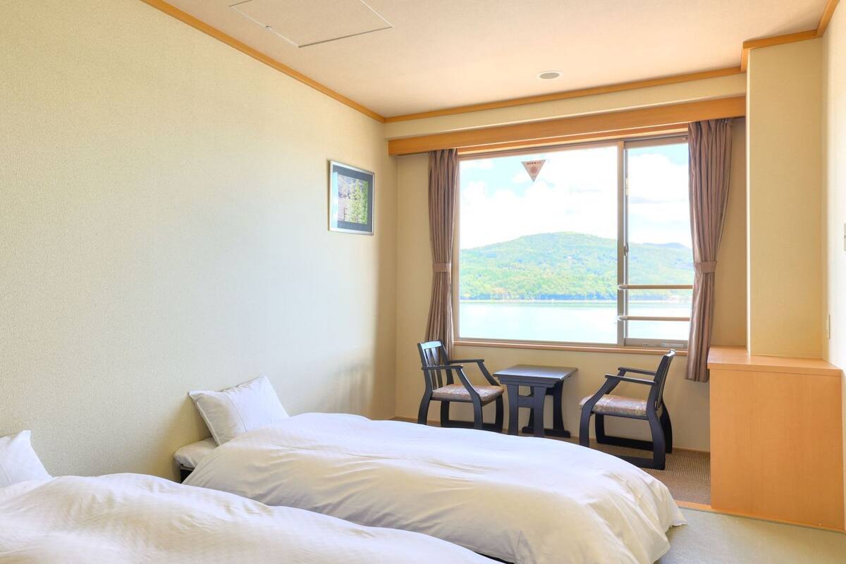 Une chambre d’hôtel avec deux lits, une petite table et une chaise près d’une fenêtre donnant sur un lac et des collines.