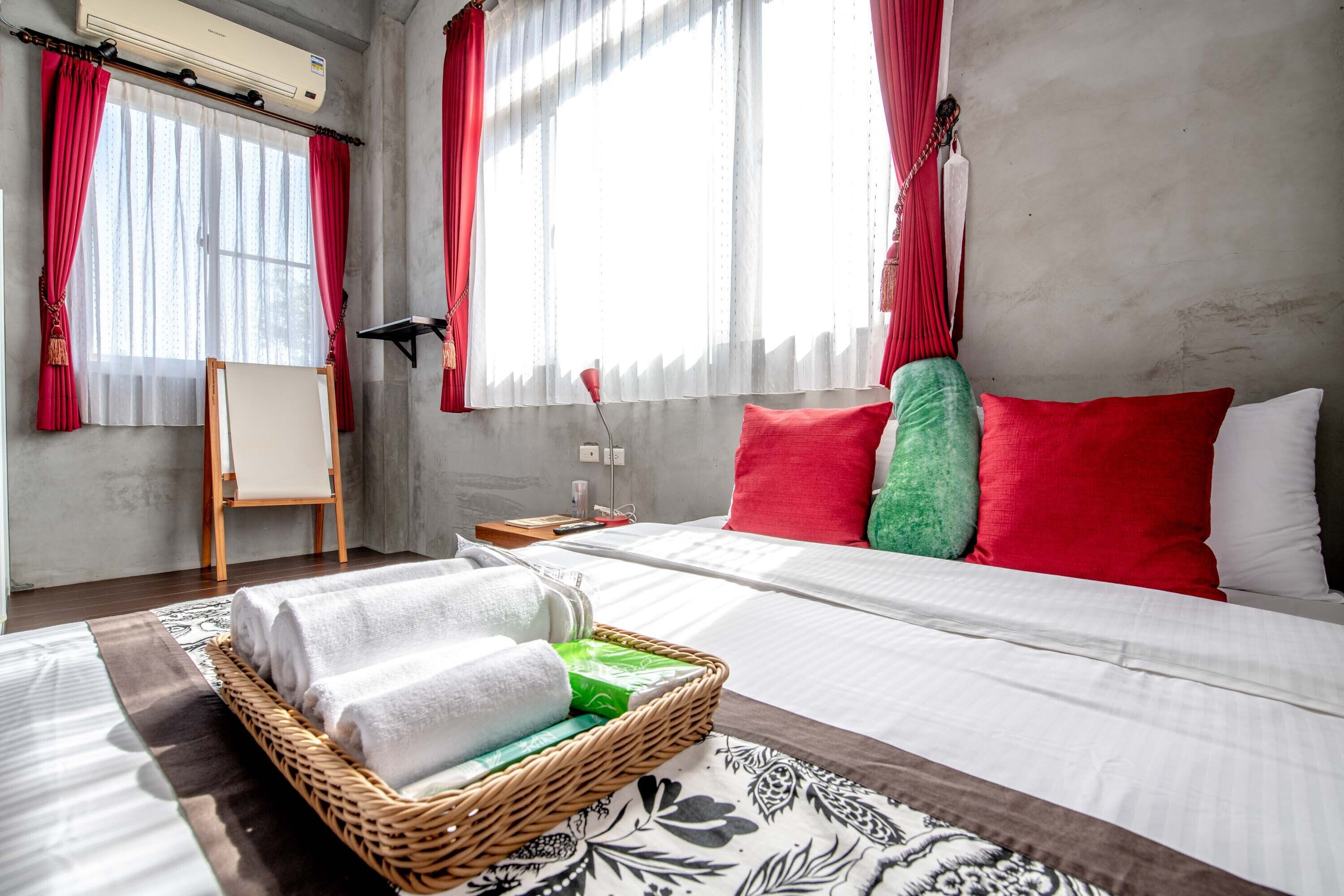 A hotel room with a bed, red and white curtains, a wicker basket with towels, and a small table with a lamp.