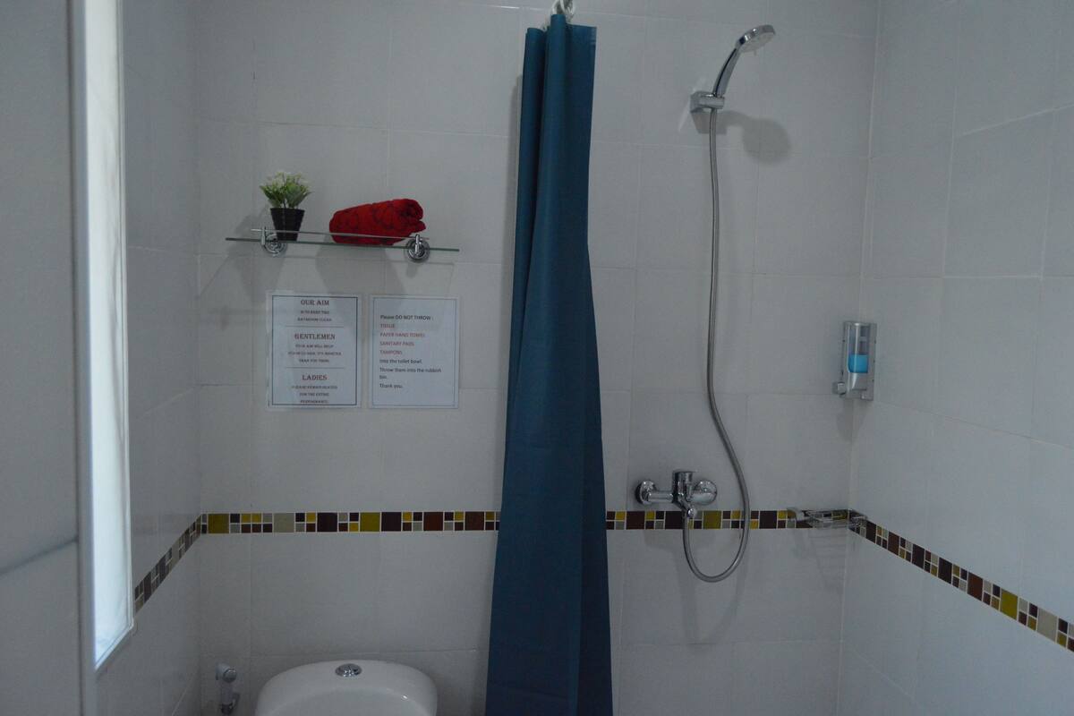 Uma casa de banho com chuveiro, sanita e lavatório. Existem sinais na parede a indicar onde colocar as toalhas e produtos de higiene.