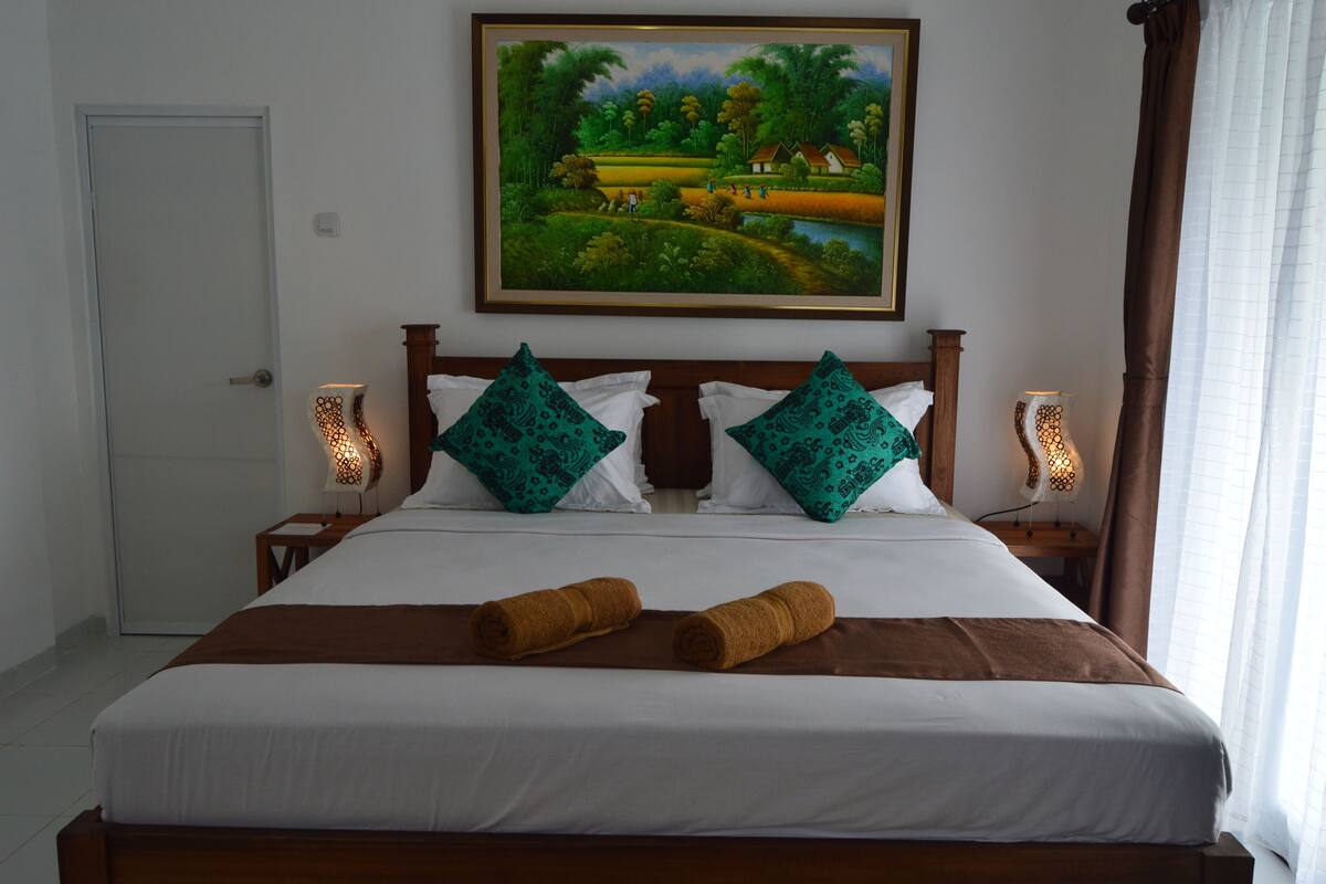 Uma cama bem arrumada com cabeceira marrom, dois travesseiros decorativos e duas toalhas enroladas.
