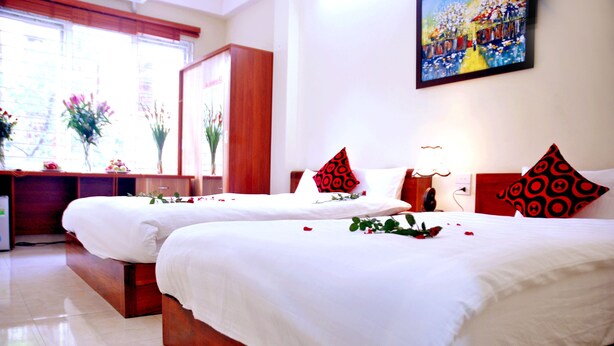 Une chambre d’hôtel avec deux lits, chacun doté de draps blancs et d’oreillers à motifs rouges. On y trouve un bureau avec une lampe et un vase rempli de fleurs.