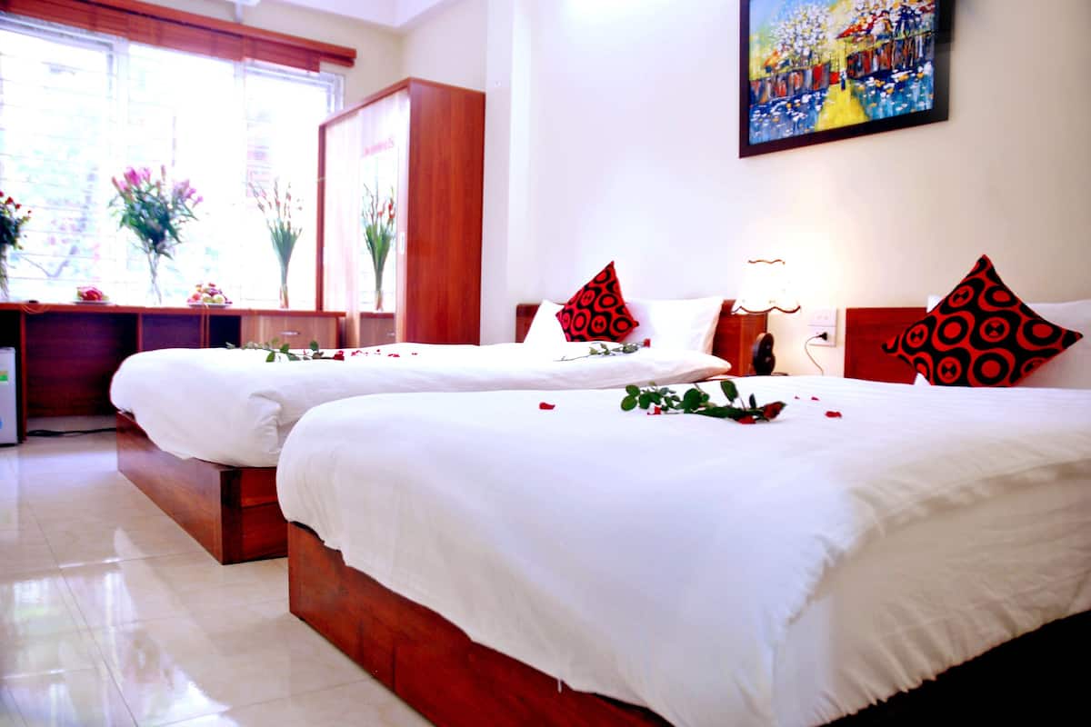 A hotel room with two beds, each with white bedding and red patterned pillows. There is a desk with a lamp and a vase with flowers.