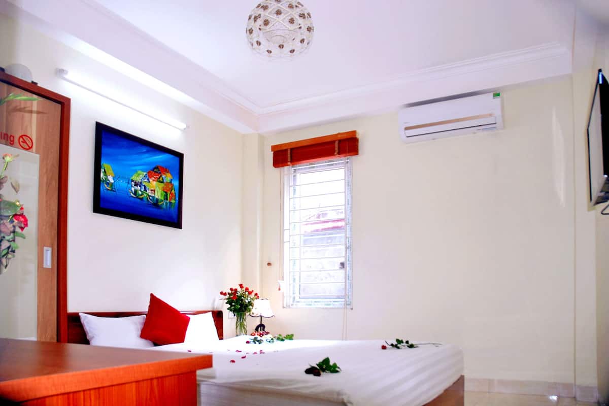 A hotel room with a bed, a desk, a window with blinds, a painting on the wall, and an air conditioning unit.