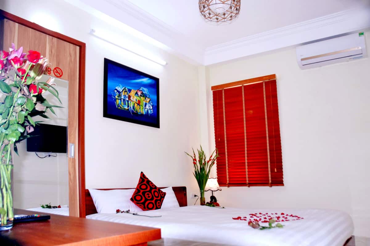 A hotel room with a bed, a TV mounted on the wall, a red window blind, and a vase with flowers on the nightstand.