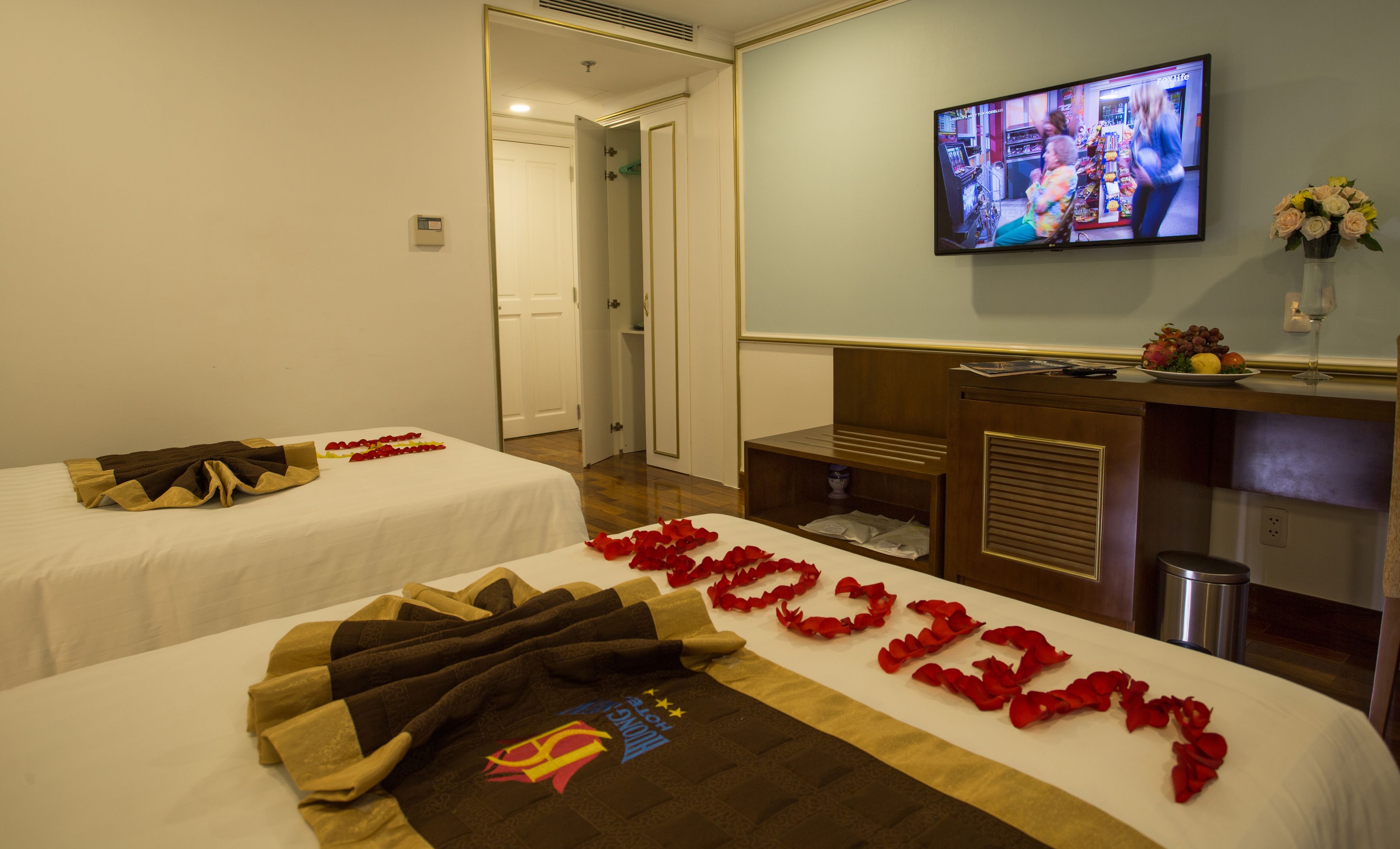 A hotel room with two beds, a TV on the wall, and a desk with a vase of flowers.