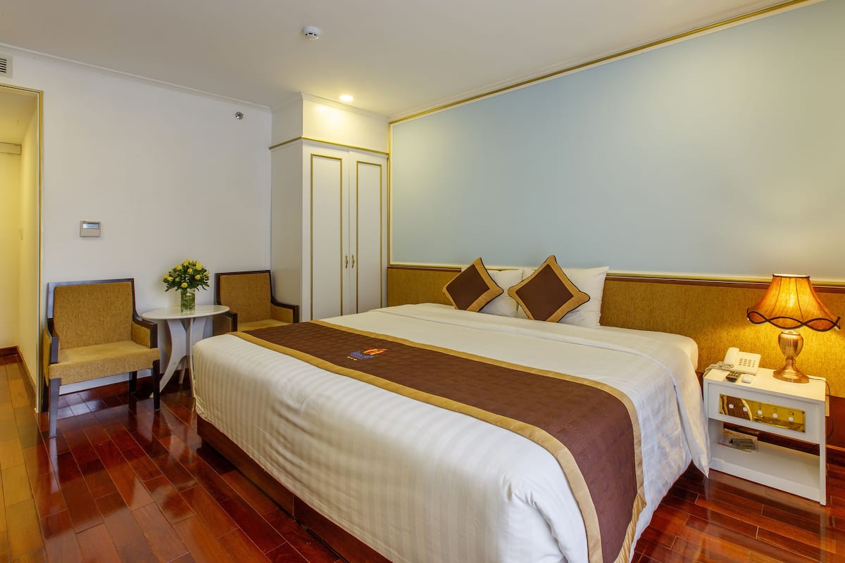 A hotel room with a large bed, a bedside table with a lamp, a chair, a small table with a vase, and a wardrobe.