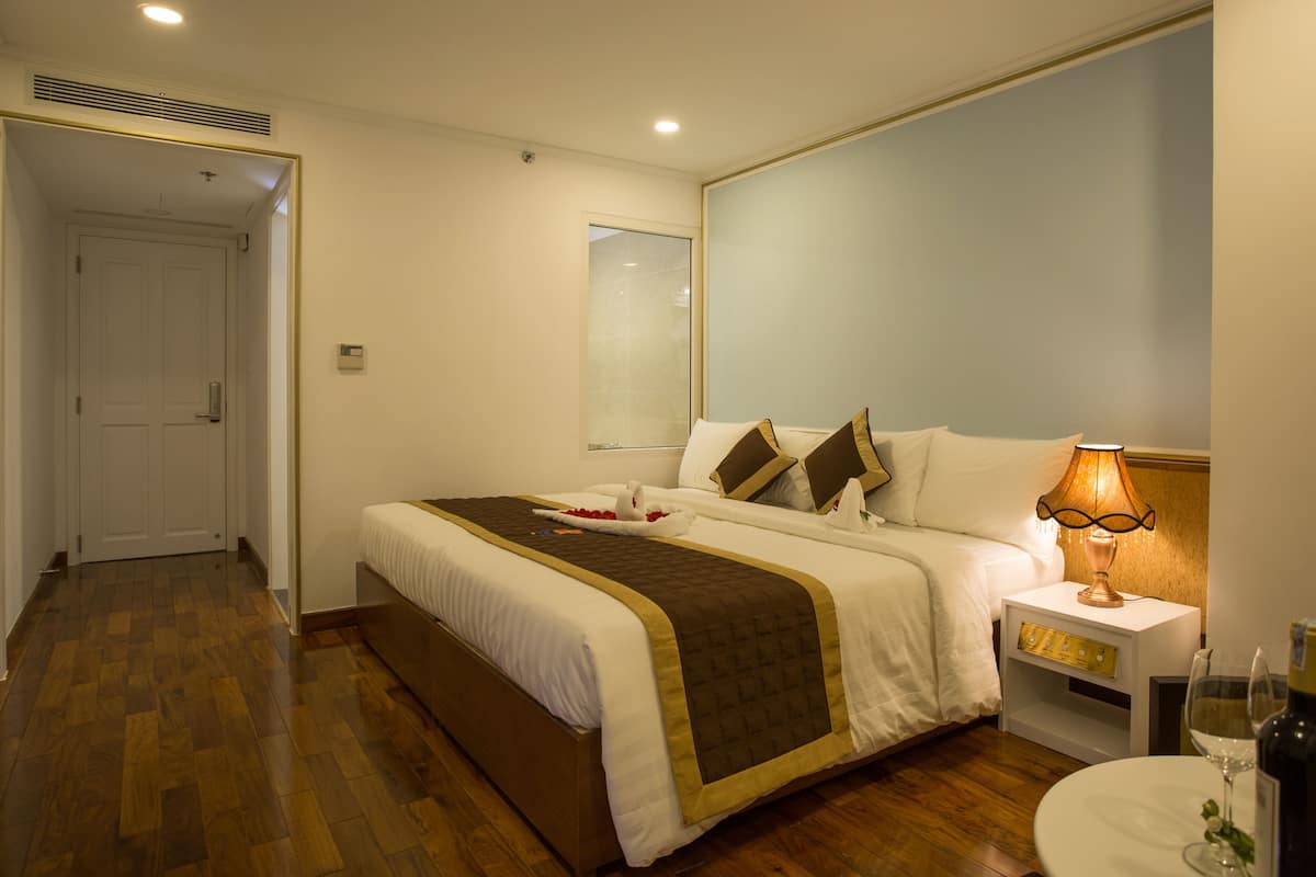 A hotel room with a large bed, a bedside table with a lamp, a wine glass, and a bottle on the right side.