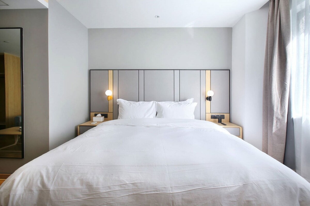 A modern hotel room with a large bed, two bedside tables, a nightstand, a mirror, and a window with curtains.